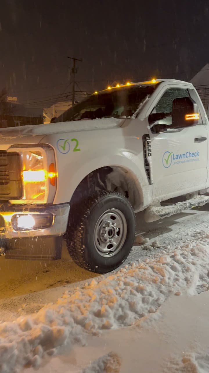 A LawnCheck plow truck clearing a snow-covered driveway at night